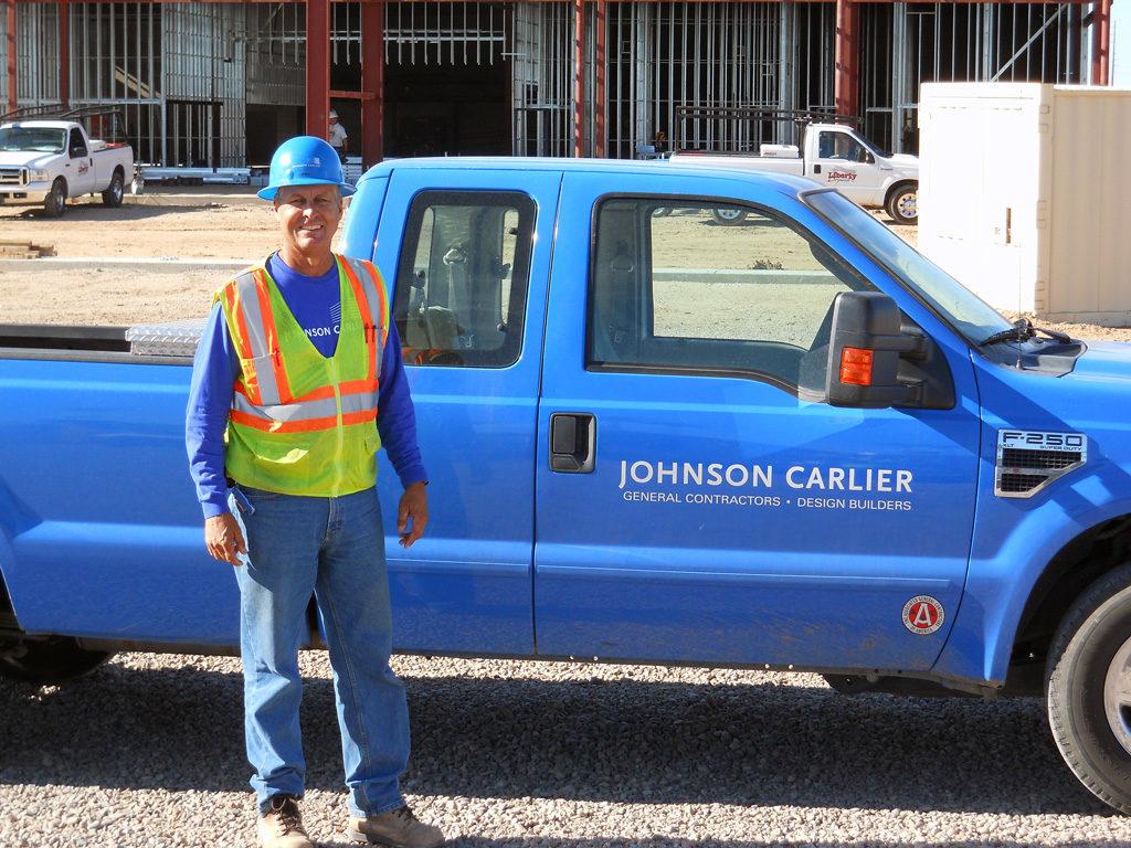 A man in construction work safety gear standing in front of a blue pickup truck with the Johnson Carlier logo on its door.